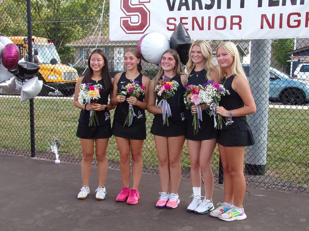 Seniors Shelbi Calligari, Lana Marsh, Madison Noonan, Julianna Vo, and Berlin Ulmer stand with their flowers in front of the banner for their senior night.