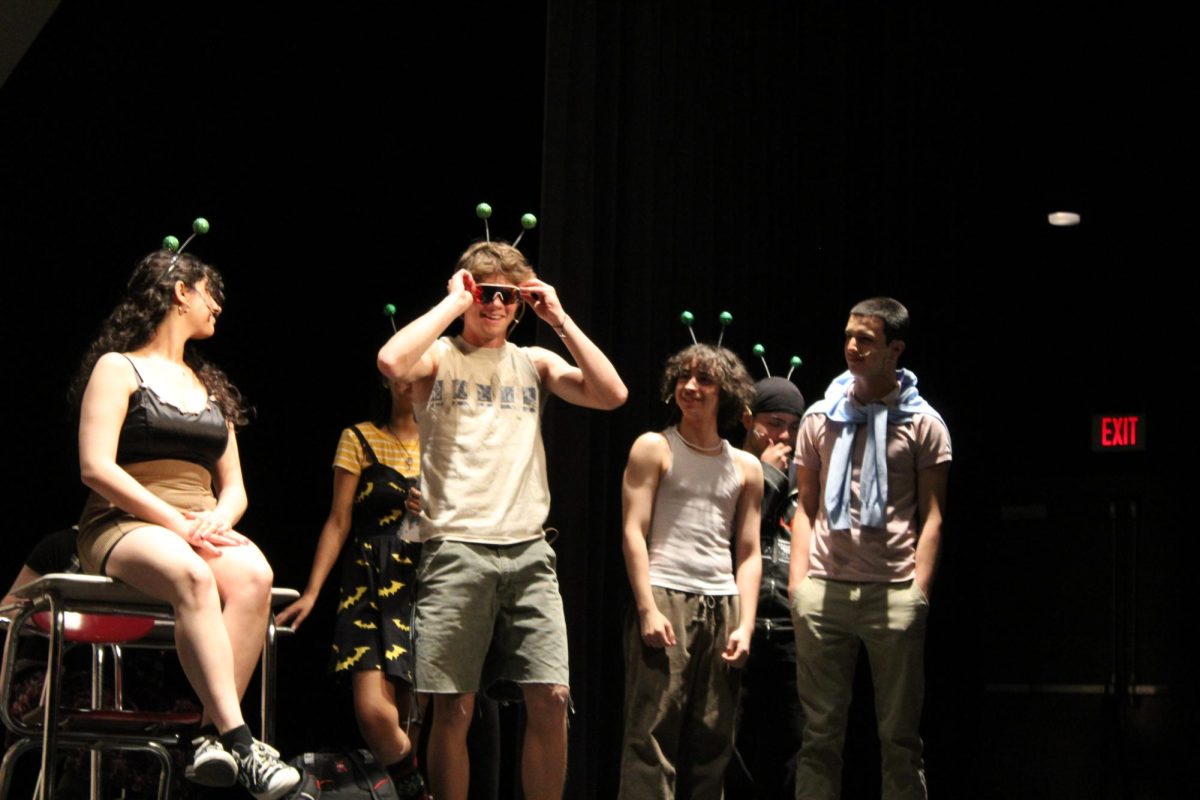 The class crowds around Ricky, played by Ben Pilcher, '25, as he fixes his sunglasses. 