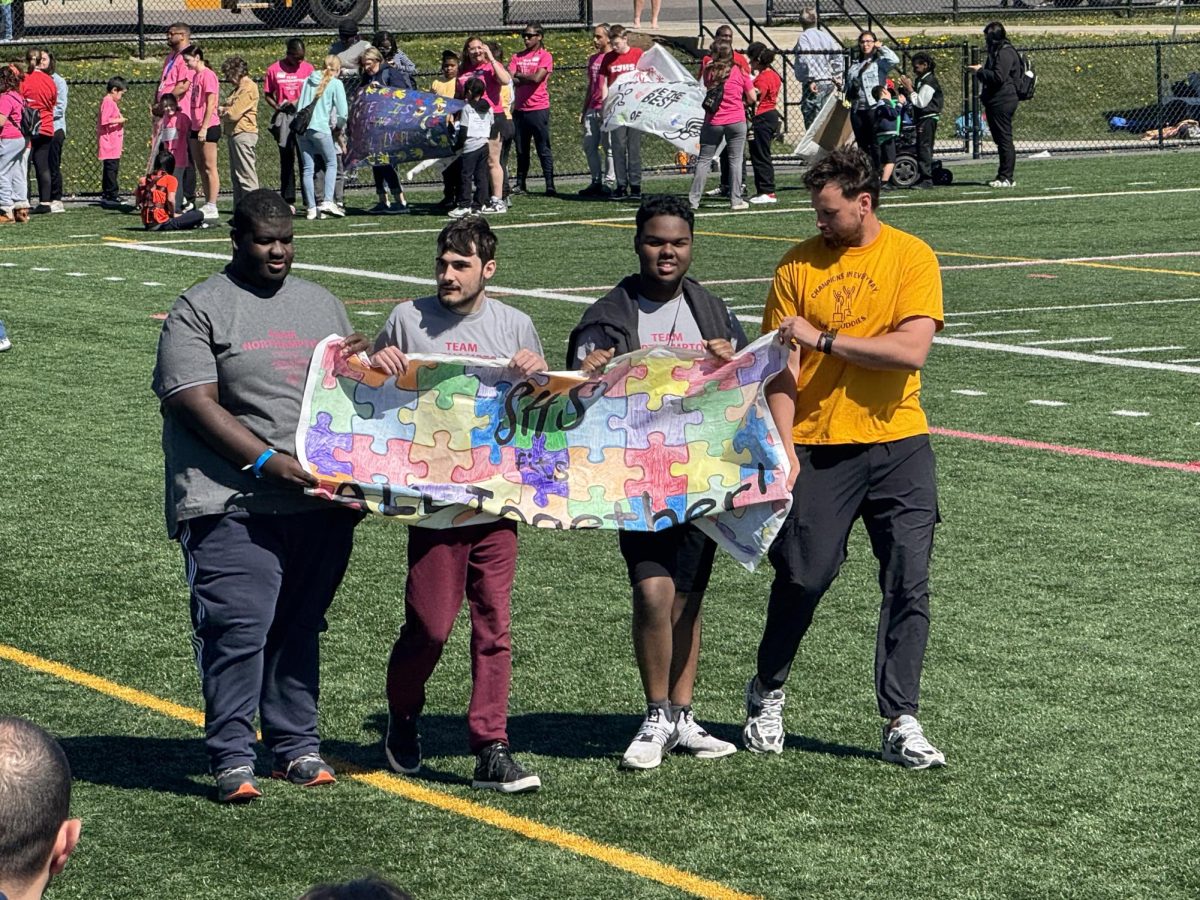 Athletes Messiah Kirkland, Mason Gearhart, and Shemar Gravesande walk the team flag on the field with Coach Cirillo.